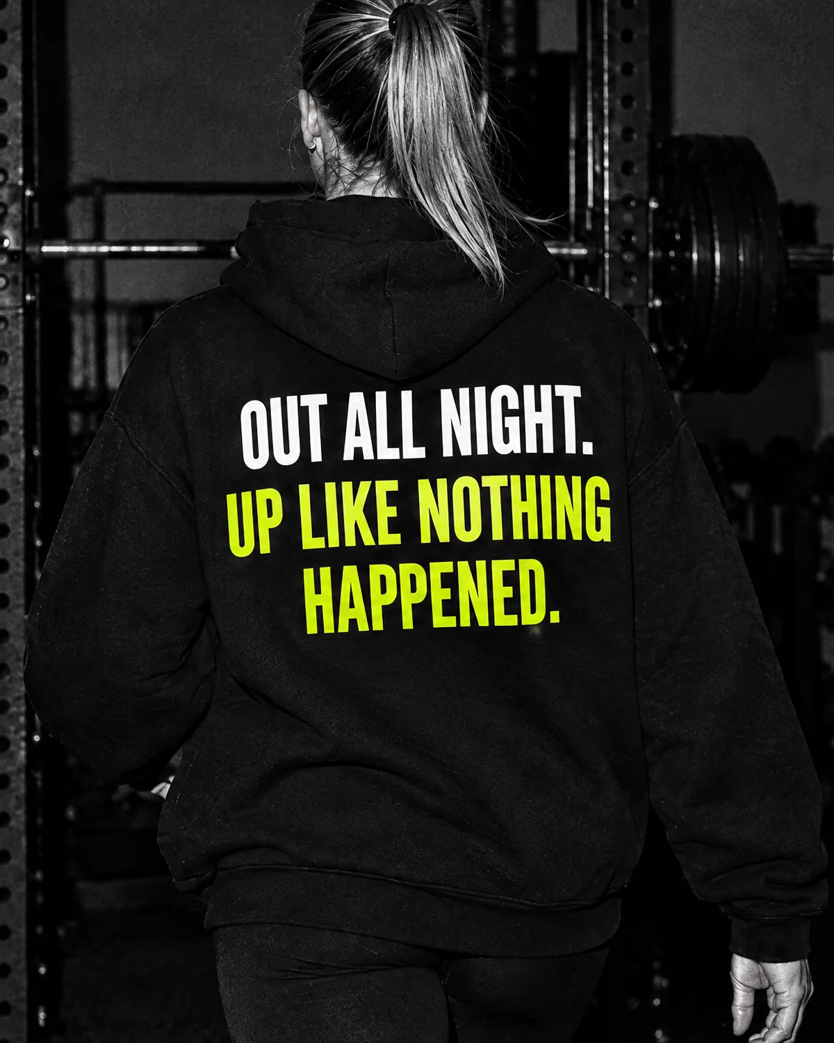 Black and white photo of a woman walking into a rooftop gym at dawn; her hoodie reads Out all night. Up like nothing happened., the second line in yellow