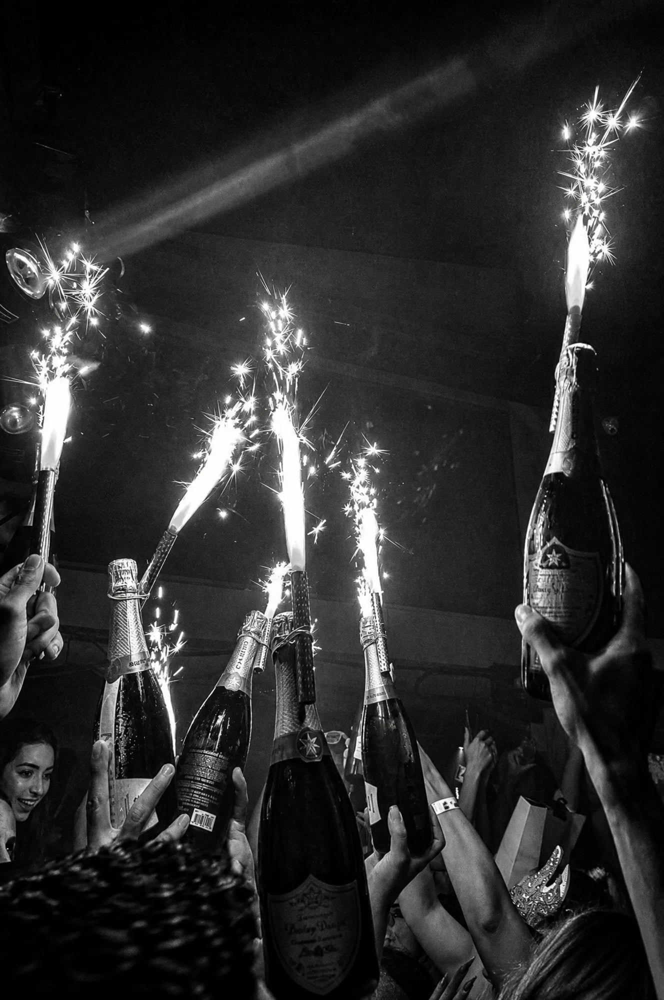 Black and white photo of hands raising sparkler-topped champagne bottles in a dark club