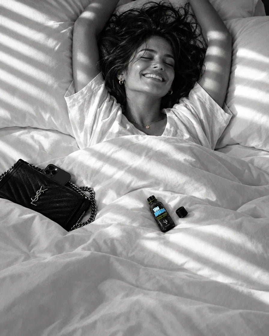 Woman waking up smiling in a sunlit bed, an empty UNHUNG bottle on the duvet beside her